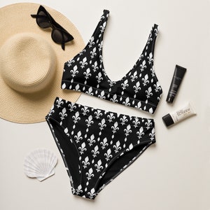 May include: Black and white swimsuit with a fleur-de-lis pattern. The swimsuit includes a bikini top and high-waisted bottoms. A straw hat, sunglasses, and sunscreen are also pictured.