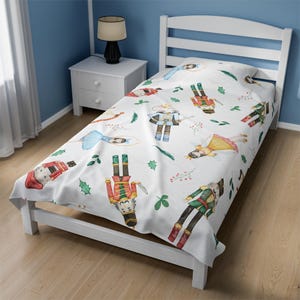 May include: White twin-sized bedspread with a Nutcracker theme. The bedspread features a repeating pattern of watercolor illustrations of Nutcracker soldiers, ballerinas, and holiday greenery.