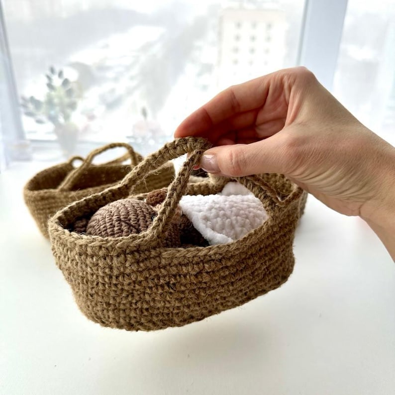 May include: A small, hand-woven brown basket with a handle, filled with a brown ball and a white crocheted item. Another similar basket is visible in the background. The basket is held by a hand.