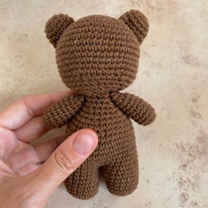 May include: A small, handmade brown crochet bear with a rounded body, small arms and legs, and two small ears. The bear is held in a person's hand. The bear is a soft toy.