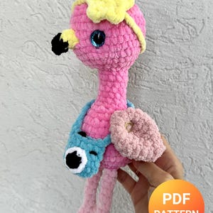 May include: A plush crochet flamingo toy made of pink yarn with a yellow star-shaped accessory on its head, a black beak, and blue plastic eyes. The flamingo wears a small blue crocheted camera bag across its body and has a light pink wing.