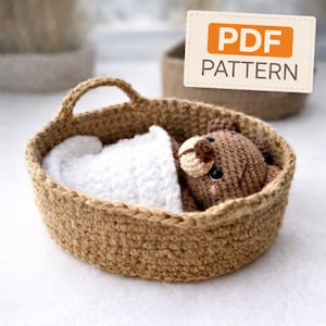 May include: A crocheted brown teddy bear nestled in a woven basket with a white blanket. The basket has a handle and is made of a light brown material. The image includes a PDF PATTERN label.