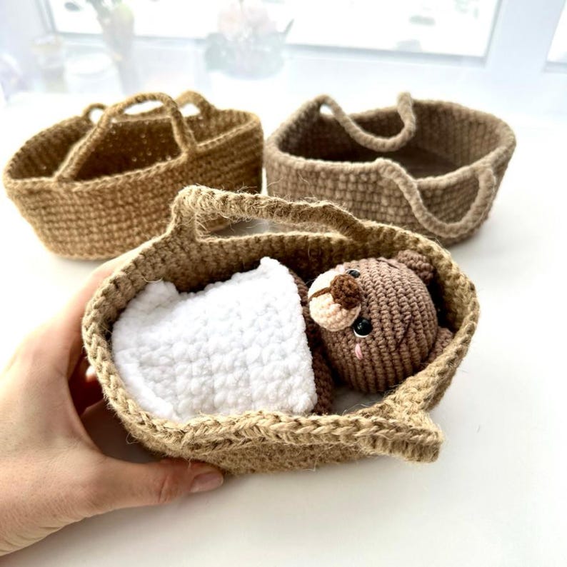 May include: A handmade crochet baby basket with a brown teddy bear and a white blanket inside. The basket is made of a natural fiber and has a handle. Two other similar baskets are in the background.