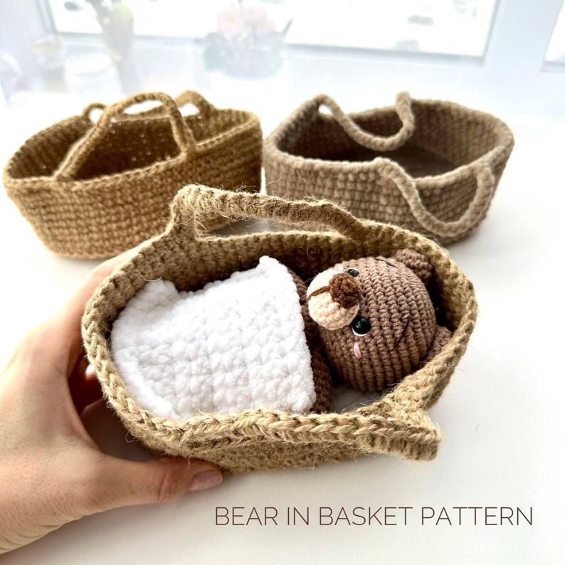 May include: A handmade crochet bear nestled in a brown basket with a white blanket. Two additional brown crochet baskets are in the background. The text "BEAR IN BASKET PATTERN" is at the bottom of the image.