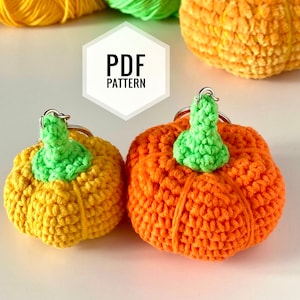 May include: Two crocheted pumpkin keychains, one orange and one yellow, with green stems. The keychains are on a white surface with the text "PDF PATTERN" in a hexagon shape.