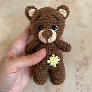 May include: A handmade brown crochet teddy bear with a light tan muzzle, ears, and belly patch. The bear has black safety eyes and a stitched mouth. The bear is being held in a hand.