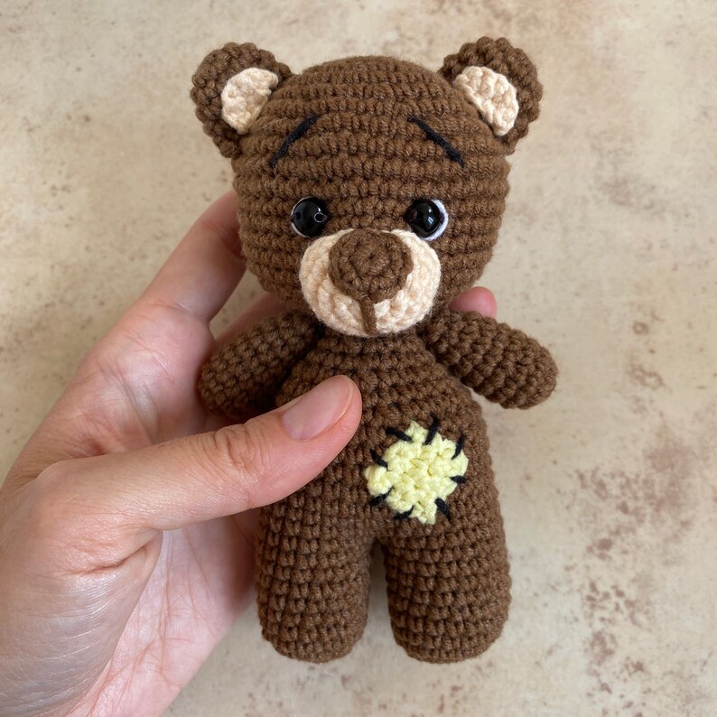 May include: A handmade brown crochet teddy bear with a light tan muzzle, ears, and belly patch. The bear has black safety eyes and a stitched mouth. The bear is being held in a hand.