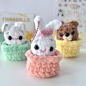 Könnte beinhalten: Drei kleine gehäkelte Plüschfiguren in pastellfarbenen Körbchen. Ein weißer Hase mit rosa Schleife im pfirsichfarbenen Korb, eine weiße Katze im mintgrünen Korb und ein brauner Bär im gelben Korb. Jede Figur hat schwarze Perlenaugen und einen rosa Mund.