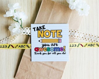 May include: A white card with the text "TAKE NOTE you are awesome! Thank you for all you do!" in colorful letters. A yellow pencil is drawn above the text. The card is on a brown paper bag with white lace and yellow ribbon with the text "123 ABC" and a red apple.
