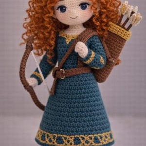 Pode incluir: Uma boneca de crochê com cabelos ruivos cacheados volumosos, vestindo um vestido azul-petróleo escuro com detalhes dourados, um cinto marrom e botas marrons. A boneca carrega uma aljava marrom com flechas e segura um arco marrom.