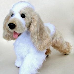 Puede incluir: Un peluche de perro Cocker Spaniel con una combinación de colores blanco y beige. El juguete tiene orejas largas y caídas, una nariz negra y una lengua rosa. El perro está de pie.