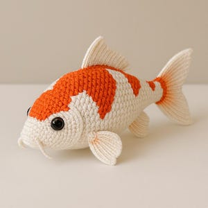 Pode incluir: Um brinquedo de peixe koi feito à mão em crochê, apresentando um corpo branco com manchas laranja vibrantes. O peixe tem uma superfície de fio texturizado, olhos de plástico preto proeminentes, barbatanas delicadas, cauda bifurcada e barbilhos.
