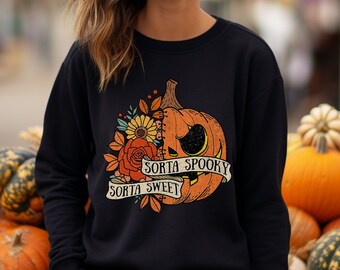 May include: A black long-sleeve sweatshirt featuring a graphic print of a pumpkin split down the middle, with one side showing a carved jack-o'-lantern face and the other side decorated with flowers. A banner reads "SORTA SPOOKY SORTA SWEET."