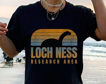 May include: A black t-shirt featuring a graphic of a silhouette of the Loch Ness monster against a sunset-colored striped background. The text reads LOCH NESS RESEARCH AREA in a bold, tan, sans-serif font centered below the graphic.