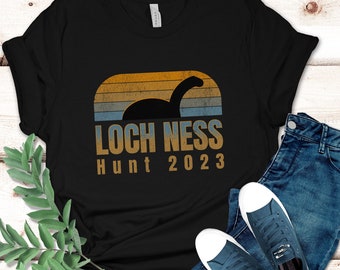May include: A black short-sleeved t-shirt featuring a graphic of the Loch Ness monster silhouette against a retro-style sunset of orange, yellow, and blue stripes. The text reads LOCH NESS Hunt 2023 in a bold, sans-serif font below the graphic.