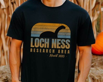 May include: A black short-sleeved t-shirt featuring a graphic of the Loch Ness monster silhouette against a sunset-colored striped arch. The text reads LOCH NESS RESEARCH AREA and Hunt 2023 in a distressed, vintage-style font and color scheme.