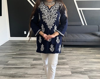 May include: A woman wearing a navy blue embroidered tunic top with white embroidery, white pants, and silver jewelry. She is standing in front of a black and white geometric wall.