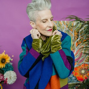 Könnte beinhalten: Eine Frau trägt einen farbenfrohen Patchwork-Mantel mit blauen, türkisfarbenen, lila und orangefarbenen Bahnen. Sie trägt auch einen grünen Samtschal und Manschetten. Der Hintergrund ist ein leuchtendes Lila.