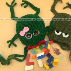 May include: Two green crocheted frog-shaped bags with googly eyes. One frog has a pink bow and the other has white eyes. Both frogs have red mouths. The bags are filled with colorful toy building blocks.