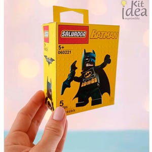 Puede incluir: Una caja amarilla estilo Lego con una figura de Batman. La caja tiene un asa y el texto "SALVADOR BATMAN" y "5+". La figura de Batman es negra con un símbolo de murciélago amarillo. La caja es sostenida por una mano.