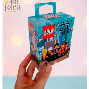 May include: A small, turquoise box with a handle, featuring the LEGO and LOLO logos, and the words "CITY cumple 6+". The box has a cityscape design and images of LEGO figures. The box is held in a hand.