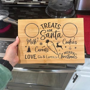May include: A wooden chopping board with a Christmas theme. It has two circles for milk and cookies, and a space for carrots. The text reads "Treats for Santa, Milk, Carrots, Cookies, Merry Christmas, Love, Gio & Lorenzo".
