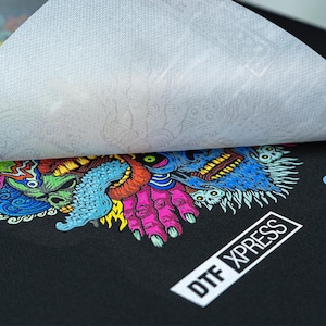 K&ouml;nnte beinhalten: Ein schwarzes T-Shirt mit einem bunten Grafikdruck eines Monsters mit einem Sch&auml;del und H&auml;nden. Der Druck ist mit einer wei&szlig;en Transferfolie abgedeckt. Der Text "DTX XPRESS" ist in der unteren rechten Ecke des Shirts gedruckt. Der Text "polyprint DTG PRINTING" ist in der oberen rechten Ecke des Shirts gedruckt.