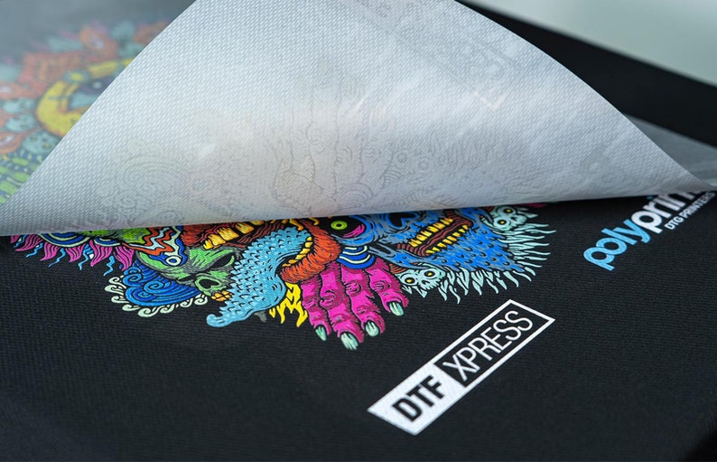 K&ouml;nnte beinhalten: Ein schwarzes T-Shirt mit einem bunten Grafikdruck eines Monsters mit einem Sch&auml;del und H&auml;nden. Der Druck ist mit einer wei&szlig;en Transferfolie abgedeckt. Der Text "DTX XPRESS" ist in der unteren rechten Ecke des Shirts gedruckt. Der Text "polyprint DTG PRINTING" ist in der oberen rechten Ecke des Shirts gedruckt.