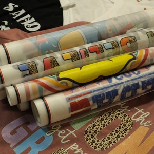 Op de afbeelding: Een stapel opgerolde hittetransfer-vinylvellen met verschillende kleurrijke ontwerpen, waaronder een regenboog, een gele smiley en een luipaardprint. De vinylvellen zijn klaar om op stof te worden geperst om aangepaste kleding te maken.