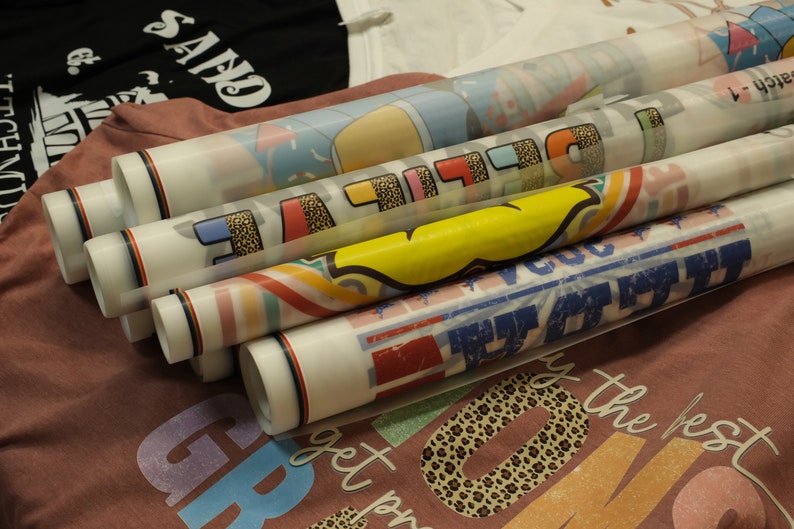 Op de afbeelding: Een stapel opgerolde hittetransfer-vinylvellen met verschillende kleurrijke ontwerpen, waaronder een regenboog, een gele smiley en een luipaardprint. De vinylvellen zijn klaar om op stof te worden geperst om aangepaste kleding te maken.