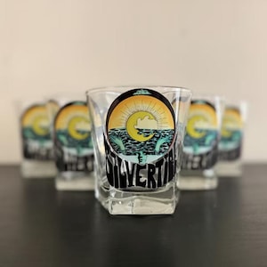 May include: Set of four clear glass shot glasses with a black and yellow graphic of a sun setting over the ocean with the text "Silvertide" printed on the front.