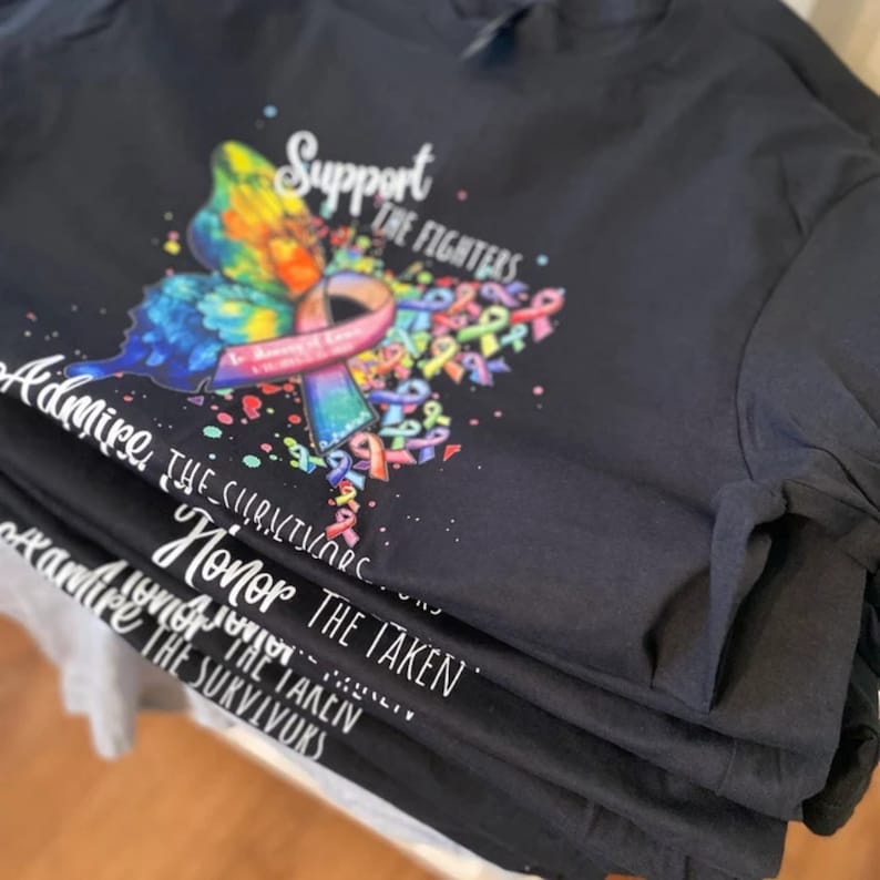 May include: A black t-shirt with a colorful butterfly design and the text "Support the Fighters, Admire the Survivors, Honor the Taken".
