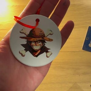 May include: White ceramic bauble with a red ribbon. The bauble features a cartoon character wearing a straw hat and a skull and crossbones design.