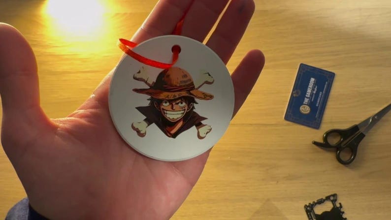 May include: White ceramic bauble with a red ribbon. The bauble features a cartoon character wearing a straw hat and a skull and crossbones design.