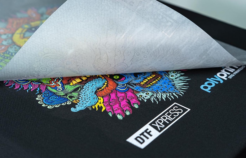 May include: A black t-shirt with a colorful graphic print of a monster with a skull and hands. The print is covered with a white transfer sheet. The text "DTX XPRESS" is printed on the bottom right corner of the shirt. The text "polyprint DTG PRINTING" is printed on the top right corner of the shirt.