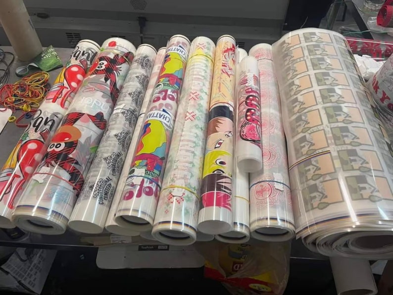 May include: A collection of rolled-up printed materials, featuring various colorful designs and patterns. The rolls are secured with rubber bands and appear to be ready for use. Some rolls have text and graphic designs.