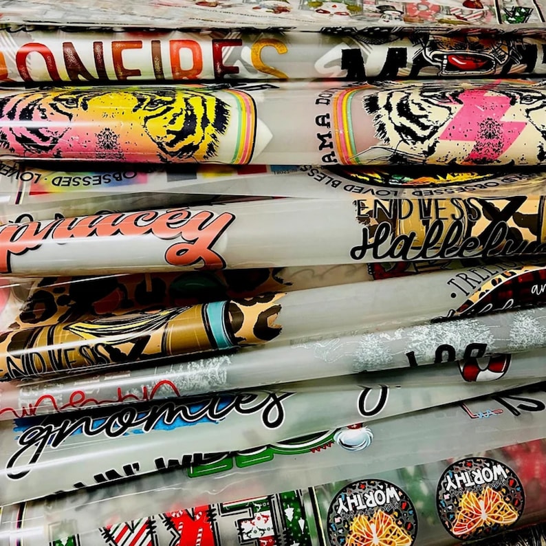 May include: A stack of clear vinyl sheets with various colorful designs printed on them. The designs include text such as "Confident", "Blessed", "Hallelujah", and "Gnomes". There are also images of tigers, butterflies, and other patterns.