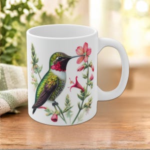 Puede incluir: Una taza de cerámica blanca con una ilustración colorida de un colibrí de plumaje verde y rojo posado cerca de flores rosas y rojas. El ave tiene un pico largo y fino y está rodeada de follaje verde y diversas flores.