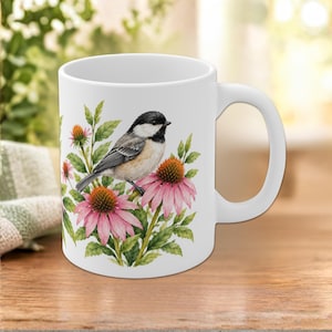 Puede incluir: Una taza de cerámica blanca con una ilustración detallada de un carbonero cabecinegro posado en un tallo entre flores de equinácea de color rosa vibrante y hojas verdes. El diseño decora la parte frontal del recipiente cilíndrico.