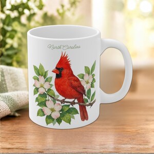 Puede incluir: Una taza de cerámica blanca con un cardenal rojo brillante posado en una rama con flores de cornejo rosa pálido y hojas verdes. El texto North Carolina está impreso en una fuente cursiva delicada sobre la ilustración del ave.