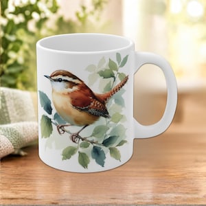 Puede incluir: Una taza de cerámica blanca con una ilustración en acuarela de un pequeño reyezuelo marrón y blanco posado en una rama con hojas verdes. El ave tiene una franja blanca distintiva sobre el ojo y una cola con patrón.