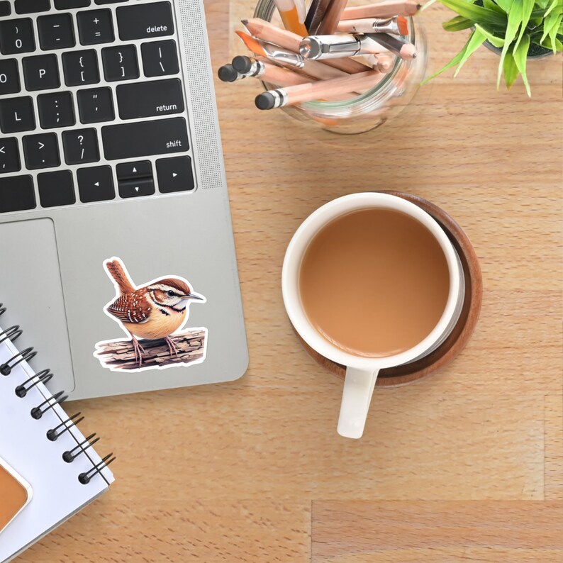Carolina Wren profile vinyl sticker adhered to a laptop on a desk. A coffee cup and casual workspace items are visible around it.