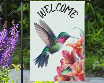 May include: A welcome garden flag with a hummingbird in flight, feeding from a cluster of orange lilies. The flag is white with black text that reads "WELCOME".