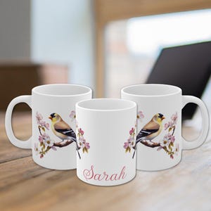 Puede incluir: Tazas de cerámica blanca con un diseño de pájaro y flores. El pájaro está posado en una rama con flores rosas. Una taza tiene el nombre "Sarah" en escritura rosa. Las tazas están sobre una superficie de madera.