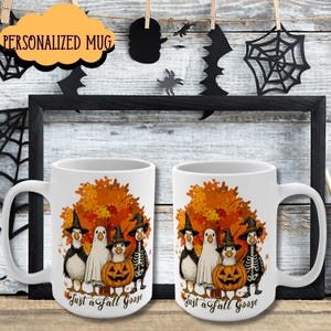 May include: Two white ceramic mugs with a Halloween-themed design. The design features geese dressed in costumes, a jack-o'-lantern, and fall foliage. The text "Just a Fall Goose" is printed on the bottom. The mugs are displayed against a backdrop of Halloween decorations.