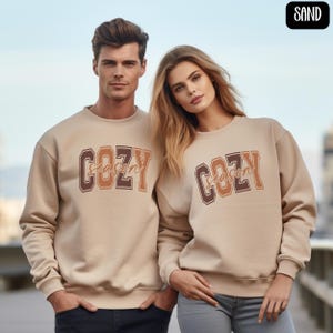 May include: Two people wearing matching sand-colored sweatshirts. The sweatshirts have the words "COZY SEASON" printed on the front in brown and orange lettering. The background is a blurred outdoor scene.