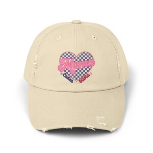 May include: A beige distressed baseball cap with a pink and blue checkered heart design. The heart has the word "Mama" in pink letters and the words "Caden" and "Olivia" in blue letters below the heart.