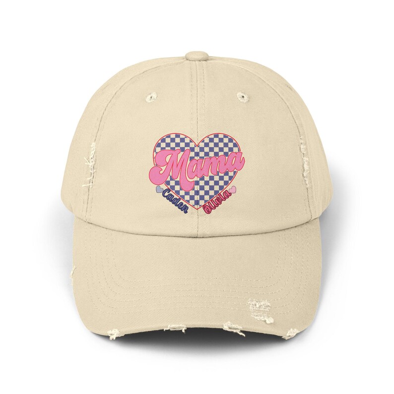 May include: A beige distressed baseball cap with a pink and blue checkered heart design. The heart has the word "Mama" in pink letters and the words "Caden" and "Olivia" in blue letters below the heart.