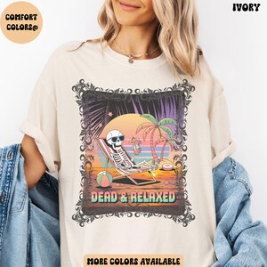 May include: Ivory-colored t-shirt with a graphic of a skeleton relaxing on a beach chair, wearing sunglasses and holding a drink. The graphic includes palm trees, a sunset, and the words "DEAD & RELAXED". The shirt has the words "COMFORT COLORS" printed on the top left.