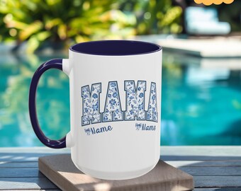May include: White ceramic mug with a navy blue rim and handle. The mug features a blue and white floral design spelling out "MAMA" with space for two names below.